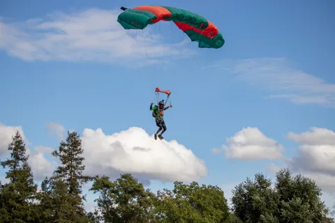 A skydiver flies a green and orange canopy above the treetops.