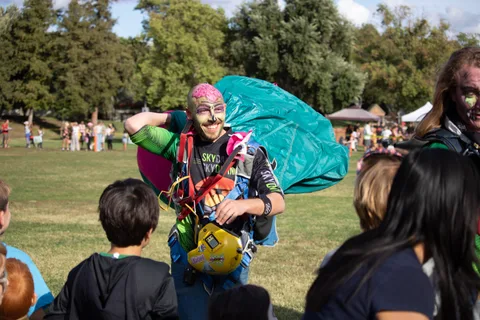 A skydiver carries a teal canopy past a row of children.