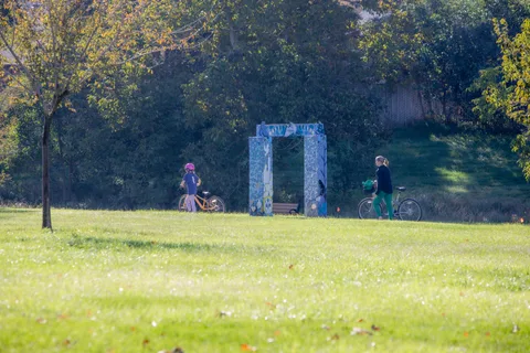 A balloon arch stands on an open lawn with a few people nearby