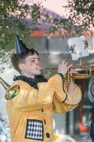 A trumpeter in a yellow harlequin costume plays in painted clown makeup.