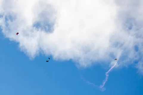 A smoke trail curls from a skydiver falling beside a cloud.