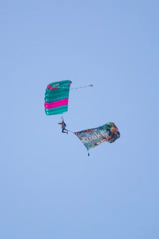 A skydiver descends under a pink and green canopy with a flag.
