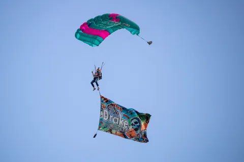 A costumed skydiver descends over Community Park trailing a Drake banner at the 2024 Zombie Bike Parade Festival.