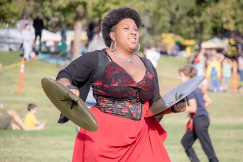 A cymbal player in a red skirt crashes her cymbals as children run past.