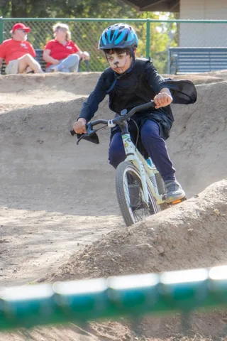 A young rider with a painted face takes the pump track on a balance bike.