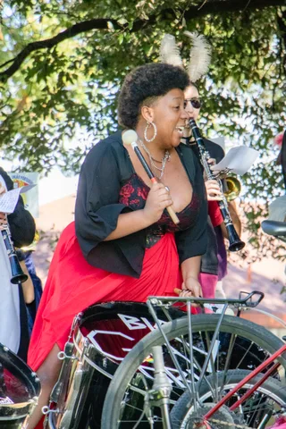 A costumed band member sings beside a bicycle under the trees.