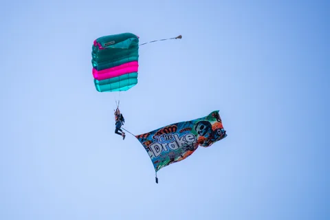 A skydiver carries a large sponsor flag under an open canopy.