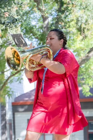A brass player in a red dress plays a baritone horn under the trees.