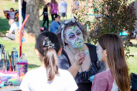 A performer in full skull face paint sits talking with visitors at the face painting table.