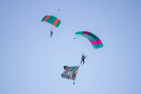 Two skydivers fly canopies with flags streaming below them.
