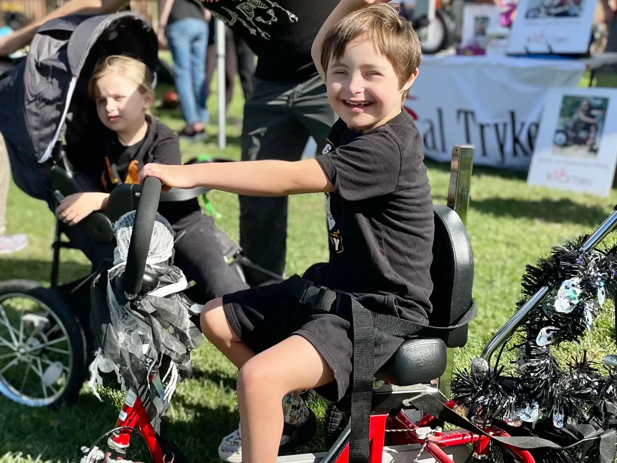 A boy laughs on a red adaptive trike on the grass at the festival, a NorCal Trykers banner behind him.
