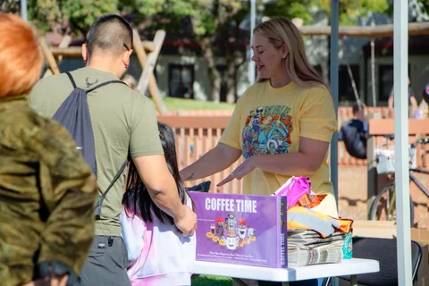 A volunteer in a festival shirt hands a prize across a giveaway table.