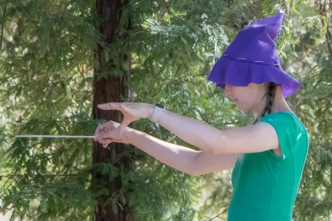 A child in a purple witch hat stretches out an arm during a game under the trees.