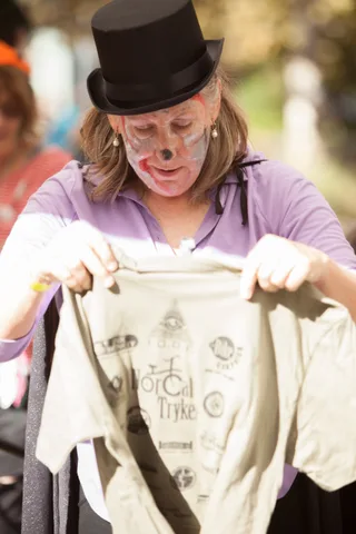 A woman in a top hat and skull makeup holds up a sponsor T-shirt printed with the NorCal Tryke logo.
