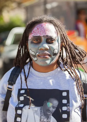 A man with intricate silver and pink face paint and dreadlocks looks at the camera.