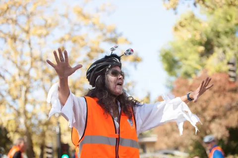 A zombie-costumed volunteer with sugar-skull antennae throws her arms wide against golden fall foliage.