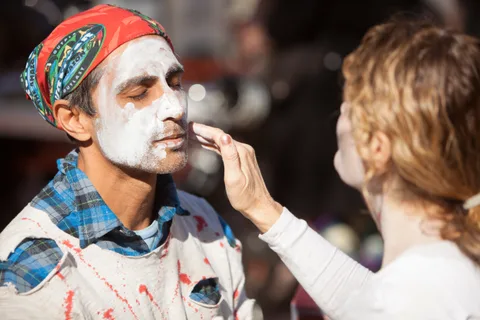 A man in a red bandana closes his eyes as white face paint is applied.