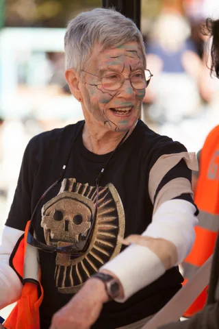 A smiling older man with gray hair has zombie face paint applied.