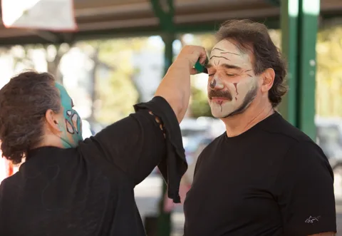 A man with a mustache closes his eyes while an artist paints skull makeup on his forehead.