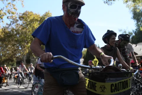 A man closes his eyes as an artist paints zombie makeup onto his face under a market awning.