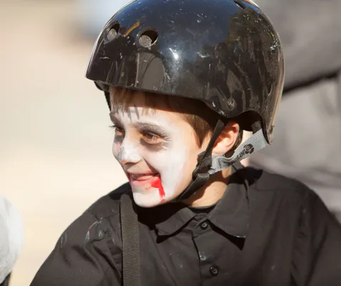 A boy in a helmet and zombie face paint smiles on his bike.