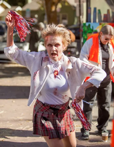 A zombie cheerleader roars mid-stride while shaking her pom-poms in the street.