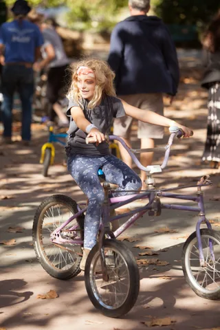 A girl with tousled hair and face paint rides a purple chopper-style bike.