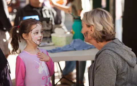 A girl has her face painted at an outdoor table while another woman looks on.
