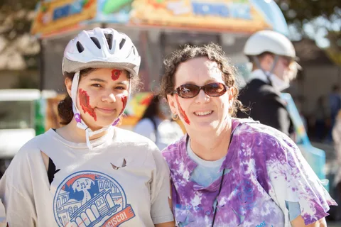 A girl in a helmet and face paint stands with a woman in sunglasses.