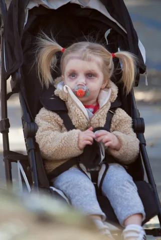 A toddler in a stroller holds a pacifier, bundled in a fuzzy jacket.