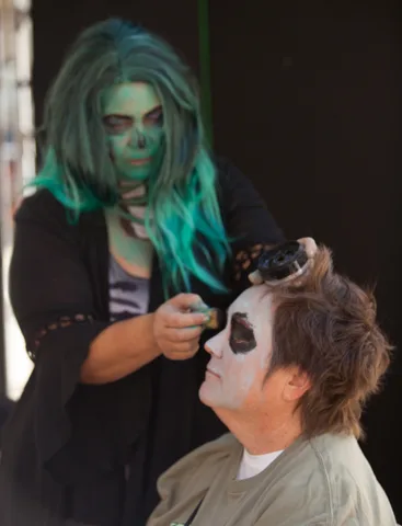 A teal-haired artist applies skull face paint to an older man with tousled hair.