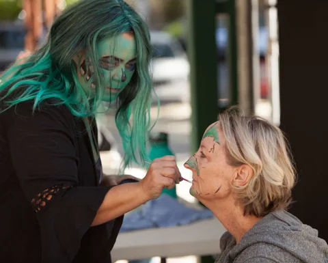 An artist with teal-streaked hair continues applying face paint to a seated woman.