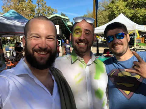 Three smiling men pose together, one with green face paint splattered on his shirt.
