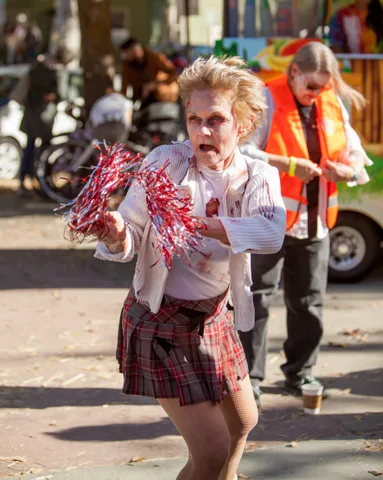 A zombie cheerleader with pom-poms strides past a volunteer in an orange vest.