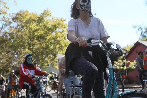 A woman in white with skull-style face paint sits on her bike among the crowd.