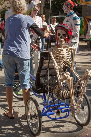A decorated skeleton in a red bike helmet rides in the basket of a blue trike among zombie-costumed riders.