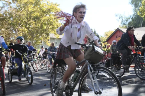 A woman in a torn zombie cheerleader costume pedals hard through the crowd with pom-poms flying.
