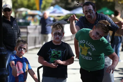 A dad in zombie makeup pretends to menace three costumed kids with a toy saw.