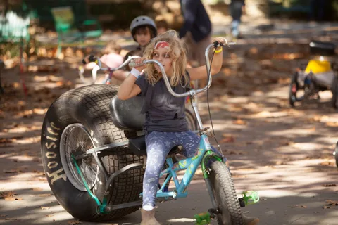 A girl rides a big-wheeled chopper trike with other kids riding nearby.