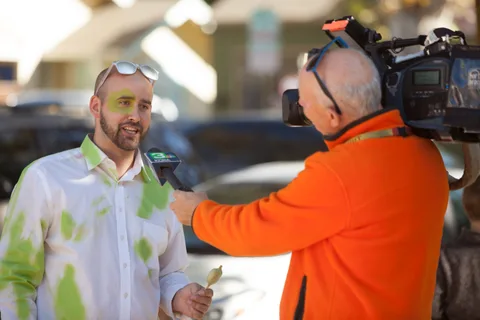 A man with green paint on his shirt is interviewed by a reporter holding a microphone.