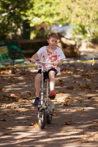 A boy with blood-stained shirt rides a bike in dappled shade.