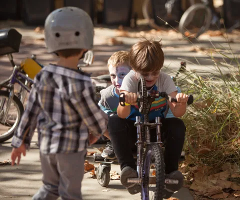Kids gather with small bikes near some shrubs.