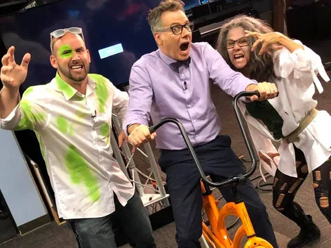 Three people laugh as green paint splatters across them at a festival booth.