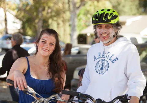 A young woman and an older man with zombie makeup and a bike helmet smile together.