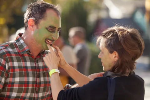 A man has green face paint applied by a woman standing close to him.