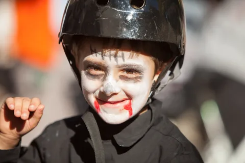A boy in a bike helmet grins with pale zombie makeup and blood dripping from his mouth.