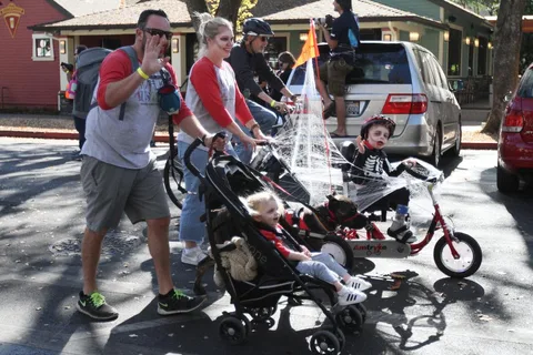 A family walks alongside a child in a skeleton costume riding an adaptive trike decorated with cobwebs.