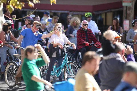 A cluster of costumed riders moves through downtown past a red building.