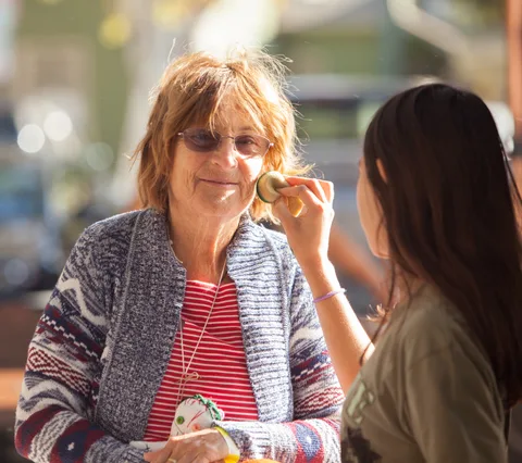 A smiling older woman has zombie makeup applied to her cheek in warm backlit sunlight.