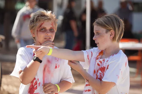 Two kids in zombie makeup and blood-stained shirts point dramatically at each other.
